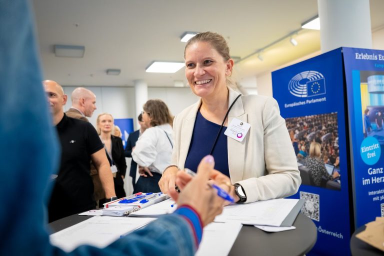 Frau im hellen Blazer lächelt beim Einchecken der Gäste, daneben ein Aufsteller des Europäischen Parlaments