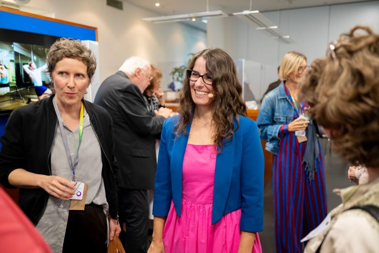 Zwei Frauen im Gespräch, eine davon in pinkem Kleid und blauem Blazer lacht