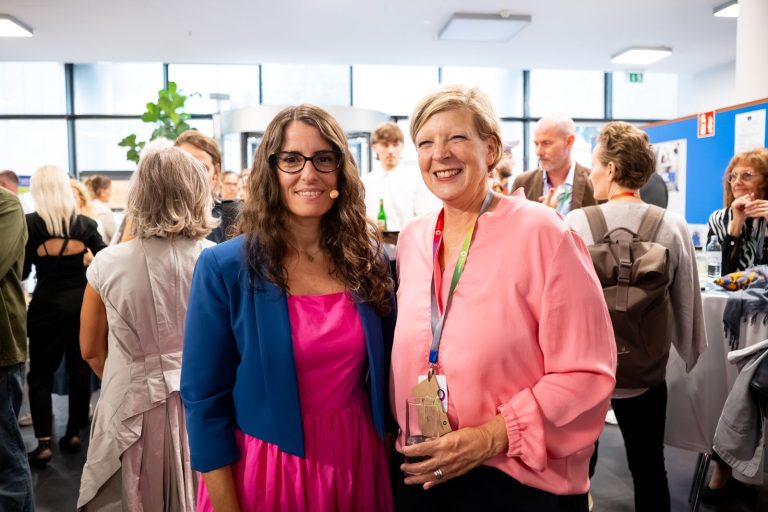 Zwei Frauen stehen lächelnd nebeneinander im Foyer, eine in pinkem Kleid mit blauem Blazer, die andere in rosa Bluse