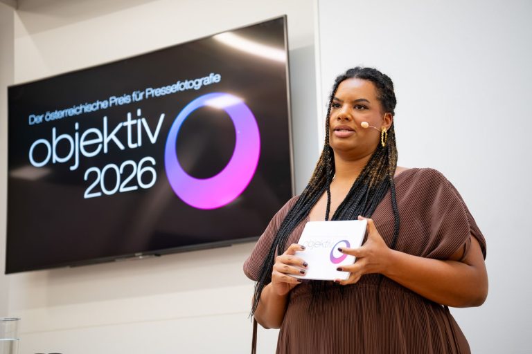 Moderatorin mit Headset spricht neben einem Bildschirm mit „objektiv 2026 – Der österreichische Preis für Pressefotografie“