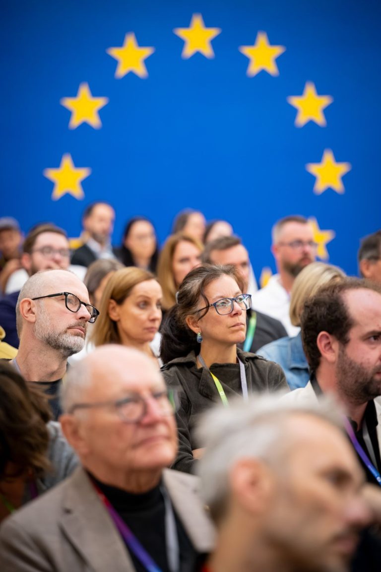 Zuhörende im Publikum vor der blauen Wand mit EU-Sternen