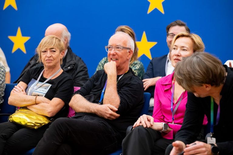 Gäste in der ersten Reihe vor dem EU-Sternenkreis hören lächelnd zu