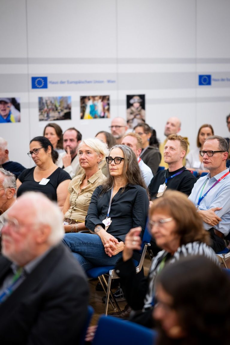 Zuhörende im Saal, an der Wand dahinter hängen Fotos