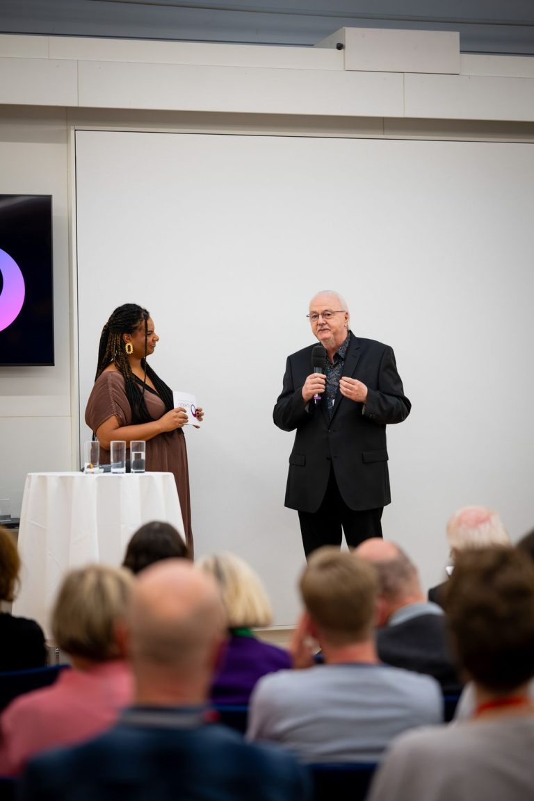 Moderatorin und ein Redner mit Mikrofon vorne im Saal vor dem Publikum