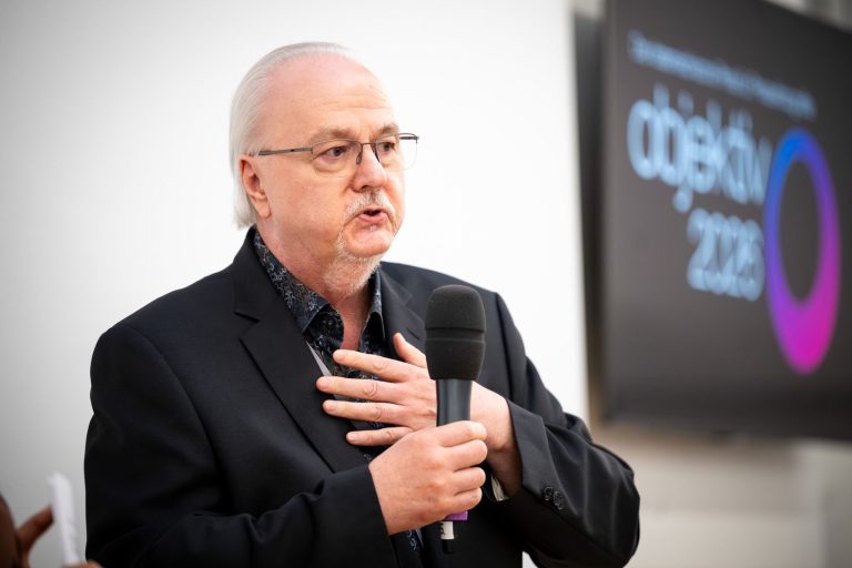 Redner mit Brille spricht ins Mikrofon, im Hintergrund das Logo „objektiv 2026“