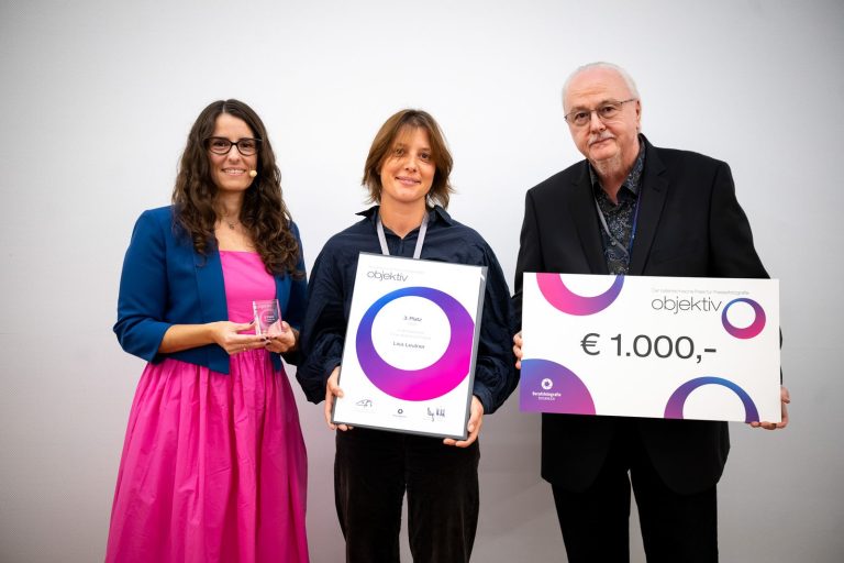 Preisübergabe Journalistische Portraits, 3. Platz: Lisa-Maria Leutner mit Urkunde, daneben die Moderatorin mit Trophäe und ein Mann mit Scheck über 1.000 Euro