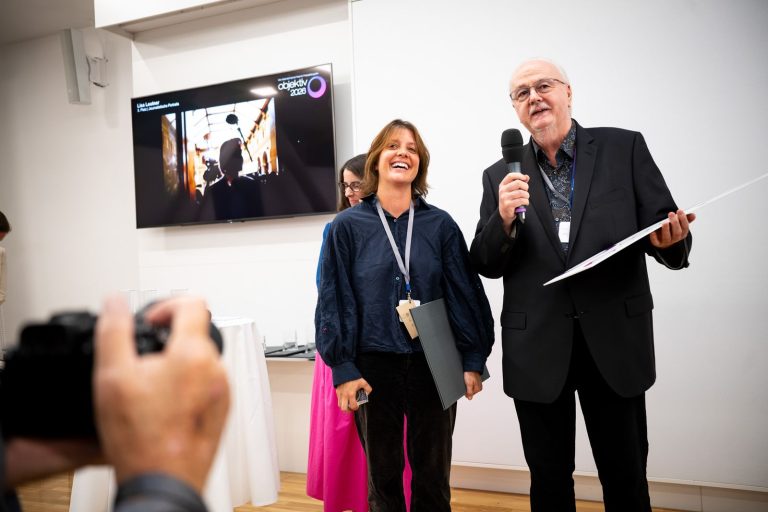 Preisträgerin lacht neben einem Redner mit Mikrofon, im Vordergrund die Kamera eines Fotografen