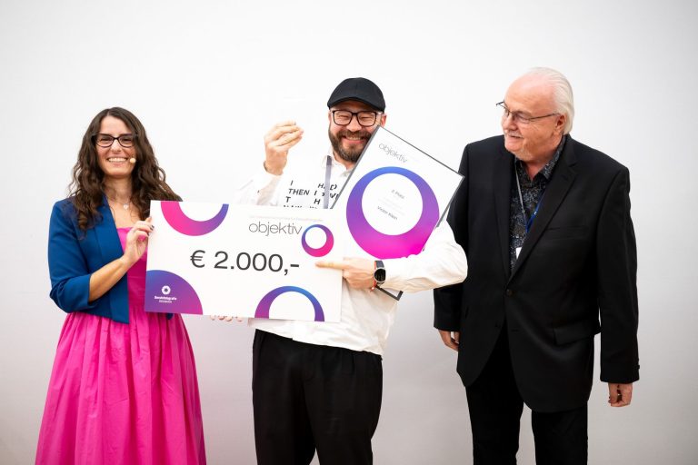 Preisübergabe Journalistische Portraits, 2. Platz: Victor Klein mit Urkunde und Scheck über 2.000 Euro zwischen der Moderatorin und einem Mann im Anzug