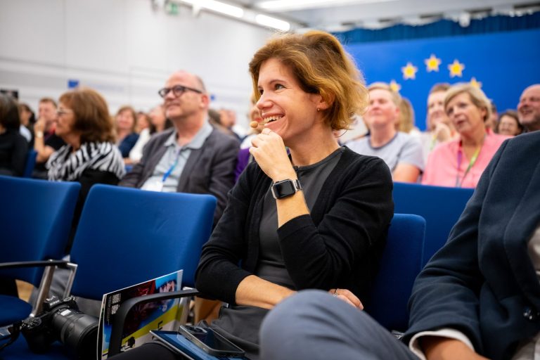 Frau im Publikum lacht, auf dem Sessel vor ihr liegt eine Kamera