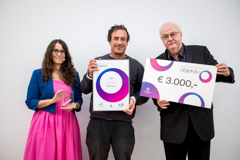 Preisübergabe Journalistische Portraits, 1. Platz: Philipp Horak mit Urkunde, daneben die Moderatorin mit Trophäe und ein Mann mit Scheck über 3.000 Euro