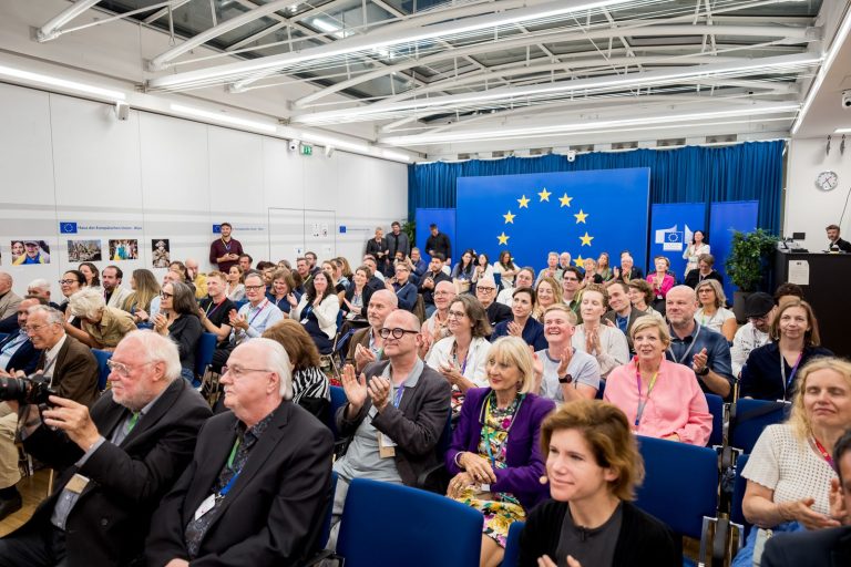 Blick über den voll besetzten Saal mit applaudierendem Publikum und EU-Sternenkreis