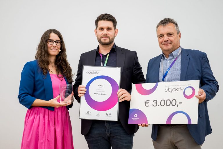 Preisübergabe Tagesgeschehen, 1. Platz: Michael Gruber mit Urkunde, daneben die Moderatorin mit Trophäe und ein Mann mit Scheck über 3.000 Euro