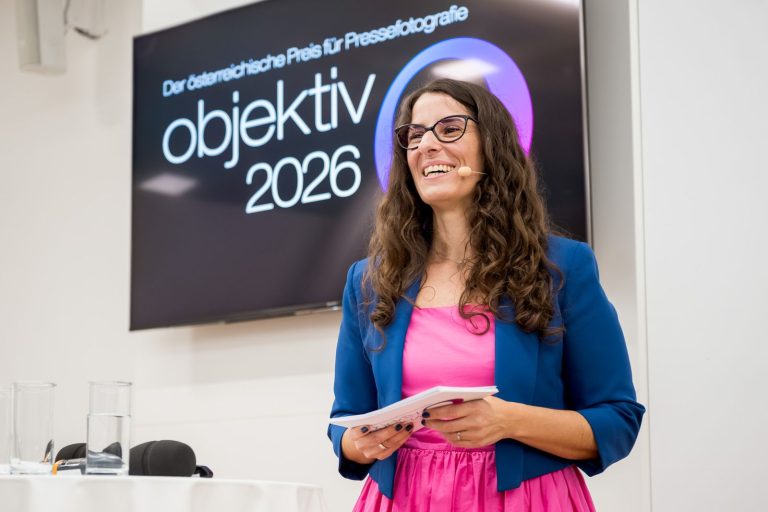 Moderatorin im pinken Kleid lacht vor dem Bildschirm mit dem Logo „objektiv 2026“