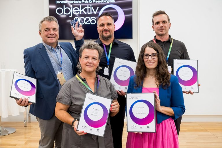 Gruppe von Preisträger:innen mit Urkunden und Trophäe vor dem Objektiv-Logo