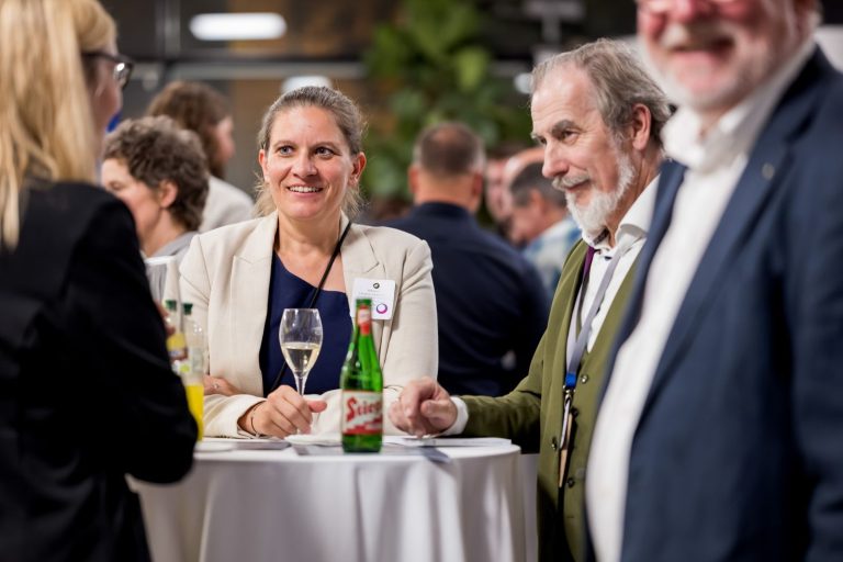 Frau im hellen Blazer und Mann mit Bart unterhalten sich an einem Stehtisch