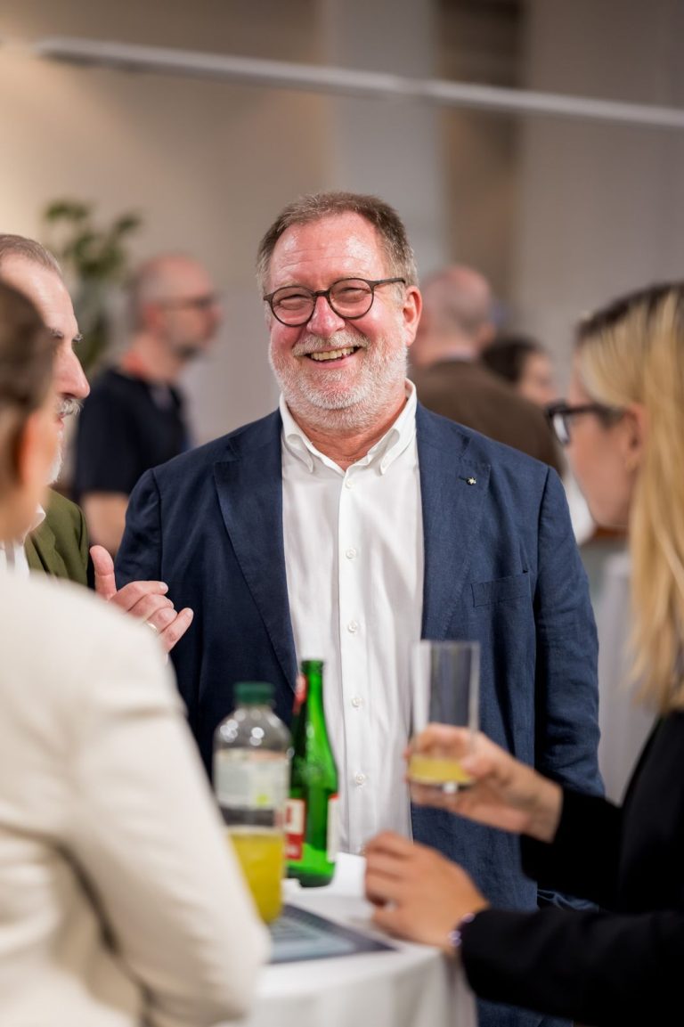 Mann mit Brille und Bart lacht im Gespräch, in der Hand ein Glas